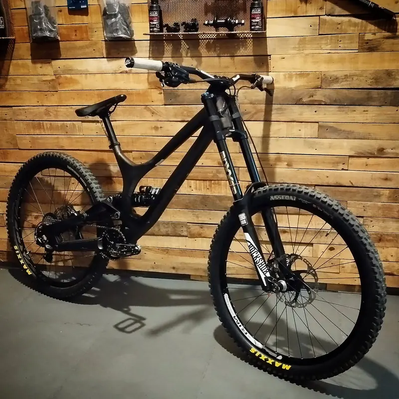 Bicicleta de downhill negra con horquilla de doble pletina, apoyada contra la pared de madera del taller.